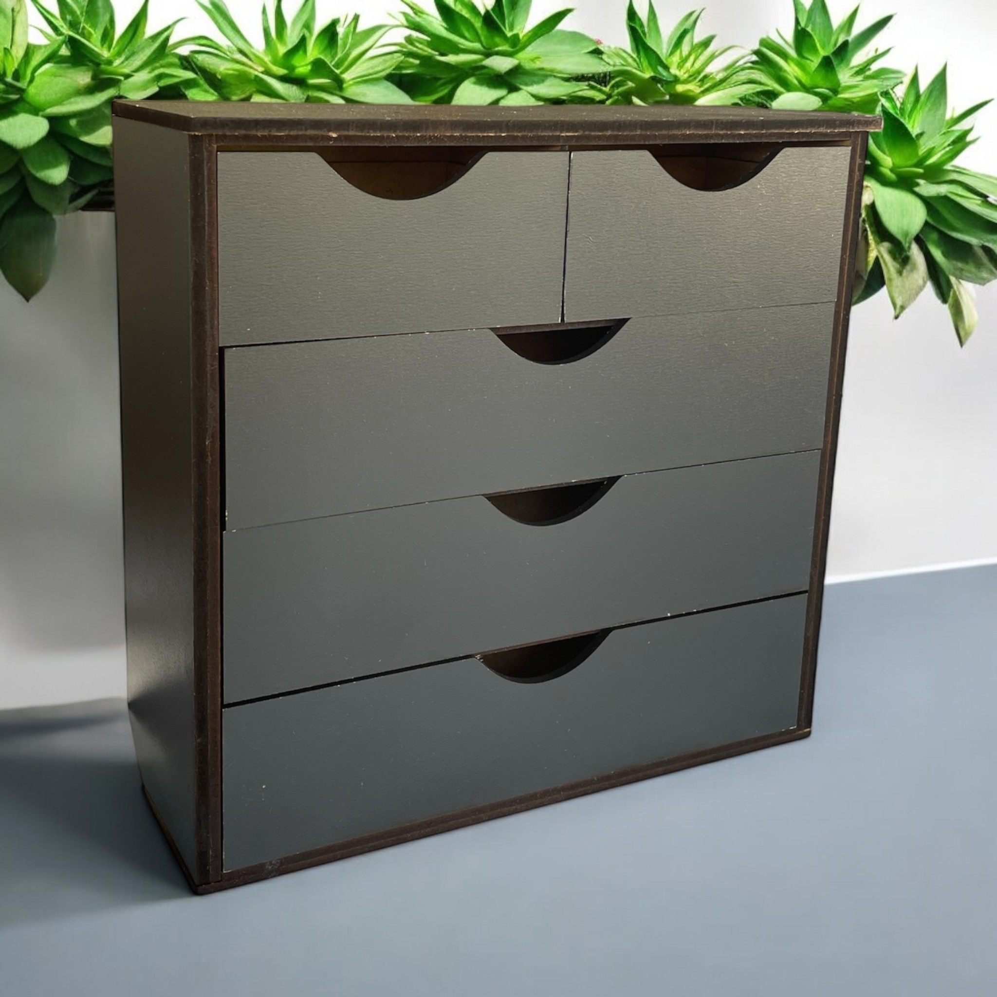 Dark brown miniature chest of drawers with cut-out handles in 1:6 scale, with a plant behind it